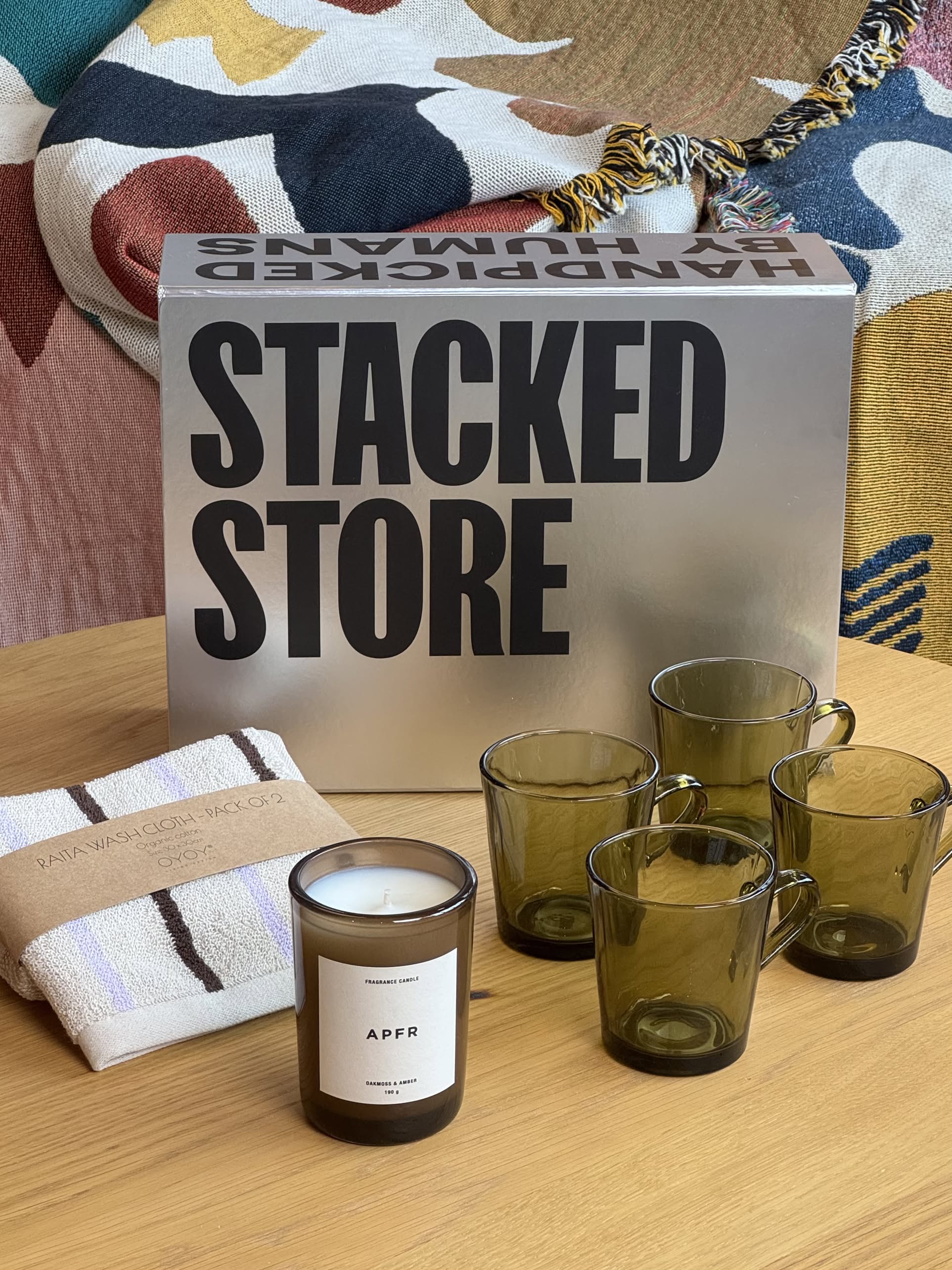 A tabletop features the Stacked Store - The New Homeowner Gift Box, four green glass cups, a white candle in a glass jar, a folded towel set by Stacked Store, and a colorful patterned blanket in the background.