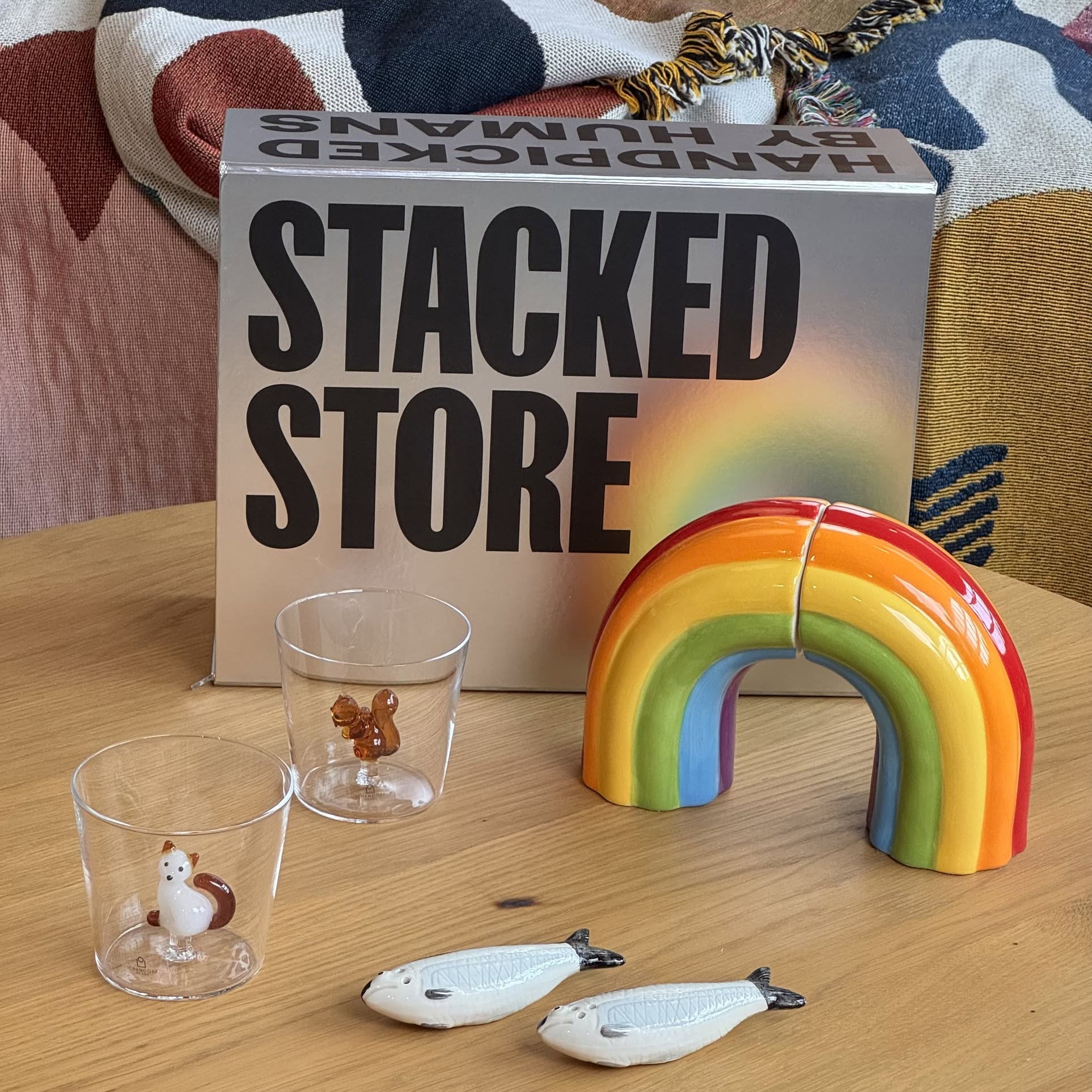 A vibrant display on a wooden table with a rainbow-shaped item, two animal-design glasses, fish-shaped pieces, and the Stacked Store The People Who Have Everything Gift Box—a unique find for design lovers. Patterned fabric sets the scene.
