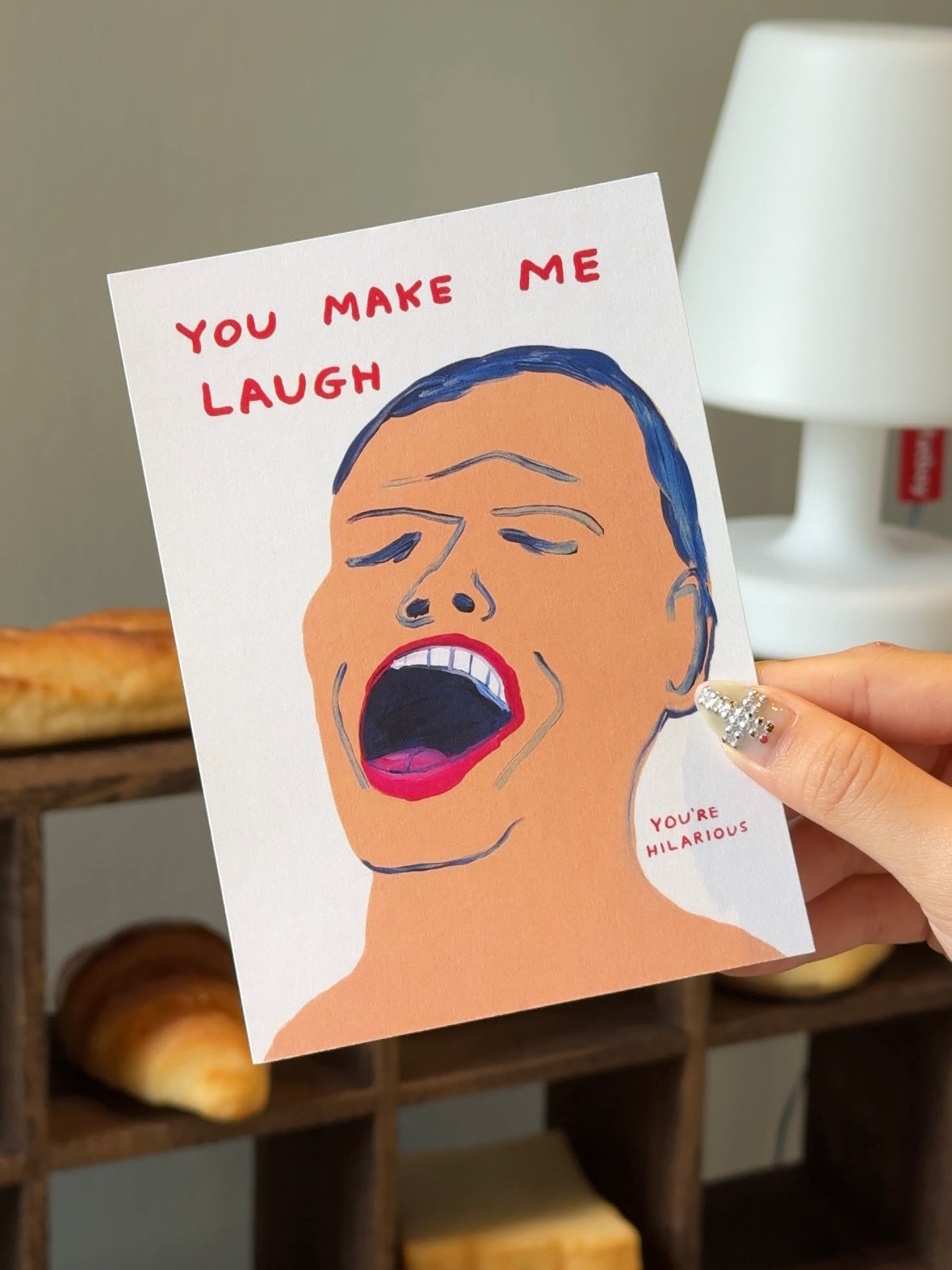 A hand holds the Stacked Store "You're Hilarious Gift Card (Physical)" featuring a laughing person, bold red text “YOU MAKE ME LAUGH” and smaller “YOU’RE HILARIOUS”—a fun, quirky pick for holiday gifting. Bread and a white lamp appear in the background.