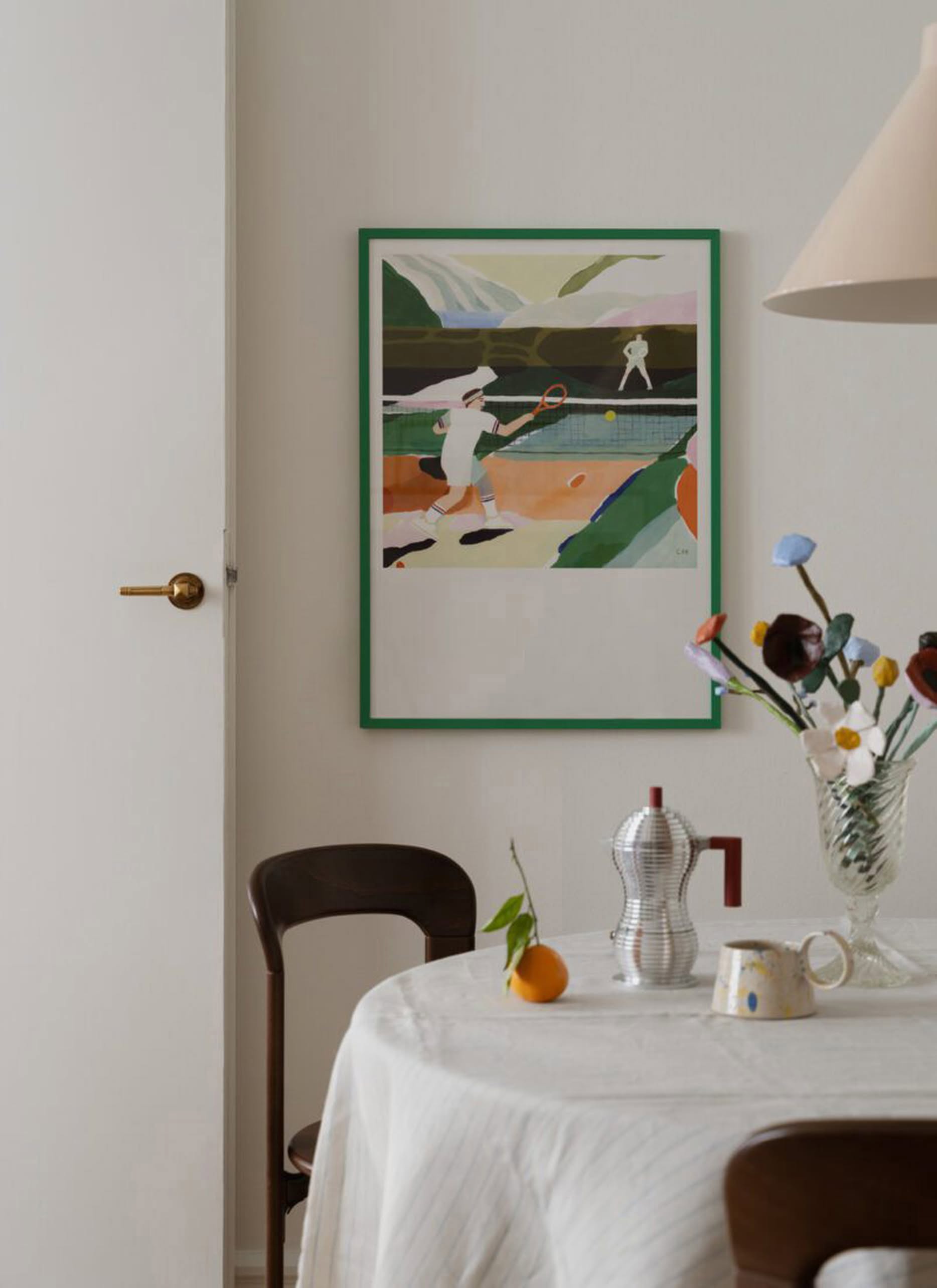 A cozy dining area features a round table with a white tablecloth, flowers in a vase, coffee maker, and an orange. On the wall hangs The Poster Club Clara Selina Bach - Tennis (30x40) poster, showcasing pastel tennis artwork.
