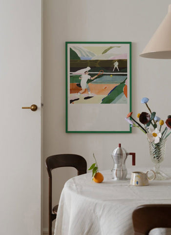 A cozy dining area features a round table with a white tablecloth, flowers in a vase, coffee maker, and an orange. On the wall hangs The Poster Club Clara Selina Bach - Tennis (30x40) poster, showcasing pastel tennis artwork.