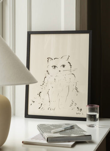 The Poster Club Oamul - Cat (30x40) poster by The Poster Club is framed and displayed on a white desk with a lamp, closed notebook, pen, and glass of water. Elegant white paneling and a dark curtain create a stylish backdrop.