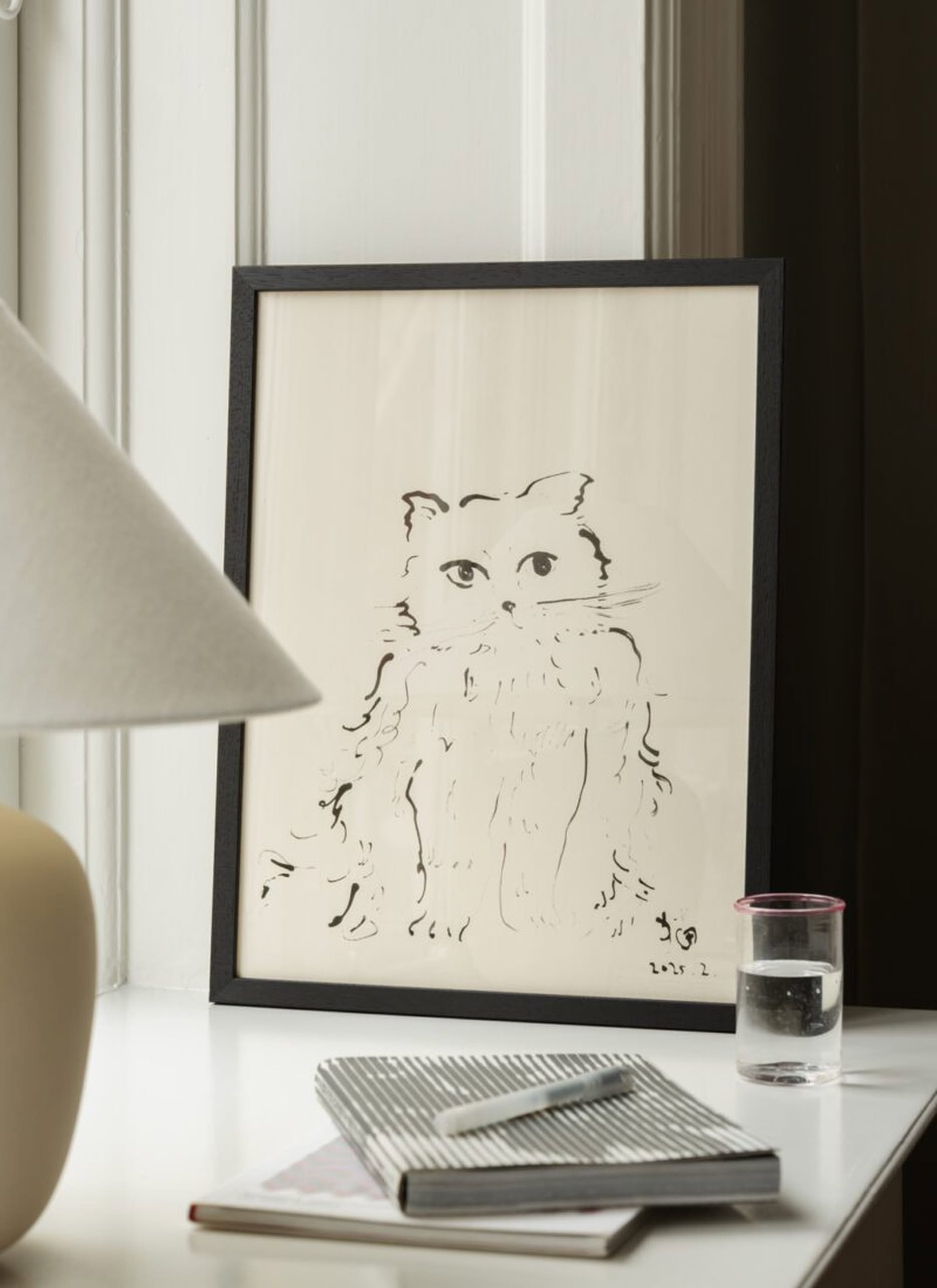 The Poster Club Oamul - Cat (50x70) poster by The Poster Club, framed in black, is displayed on a white table with a notepad, pen, glass of water, and beige lamp. The setting has white and dark brown walls in the background.