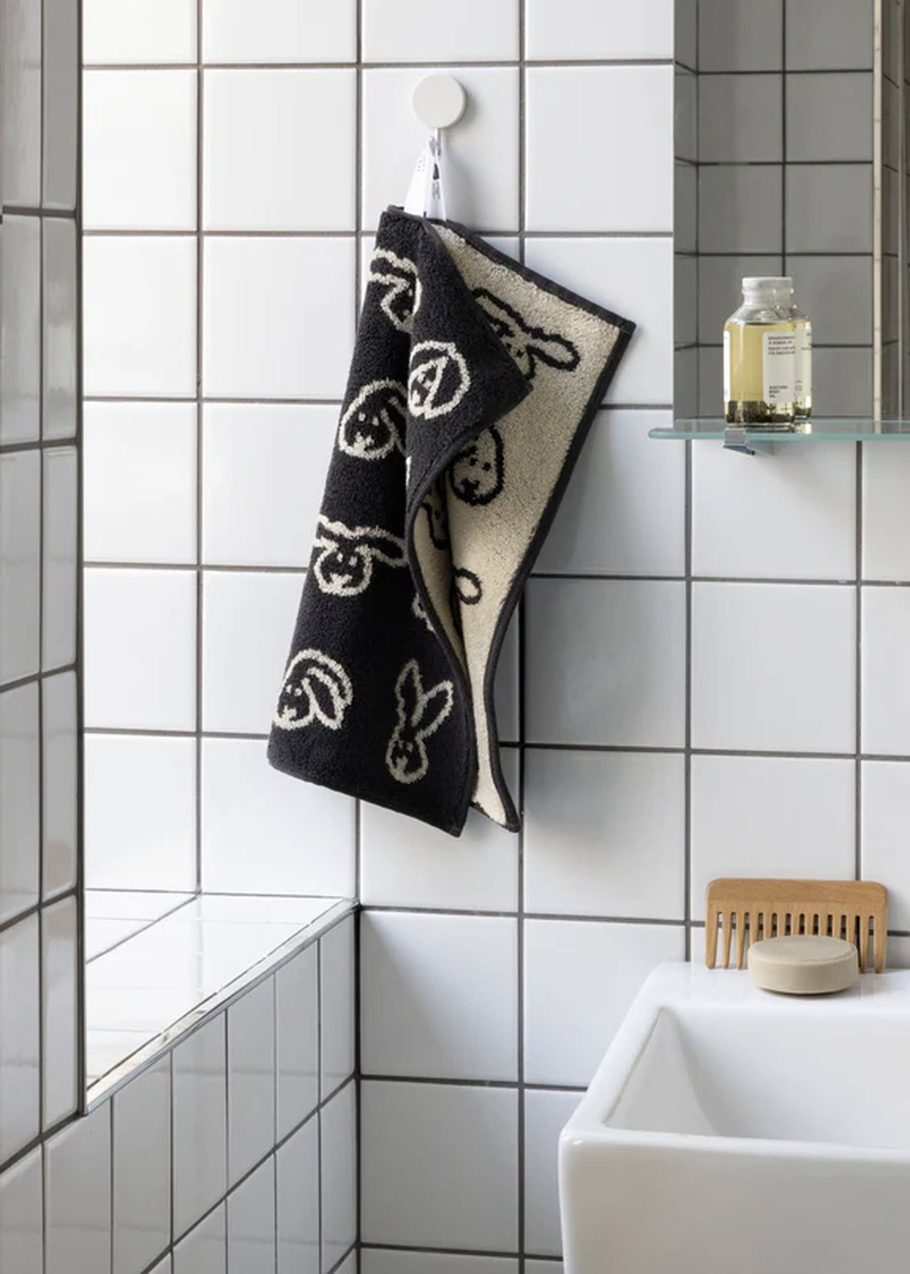 The WARMGREY TAIL Bunny Bunny Hand Towel, featuring a black and white abstract face pattern, hangs on a hook in a tiled bathroom next to a mirrored shelf with a bottle and brush near the sink.