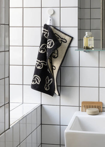 The WARMGREY TAIL Bunny Bunny Hand Towel, featuring a black and white abstract face pattern, hangs on a hook in a tiled bathroom next to a mirrored shelf with a bottle and brush near the sink.