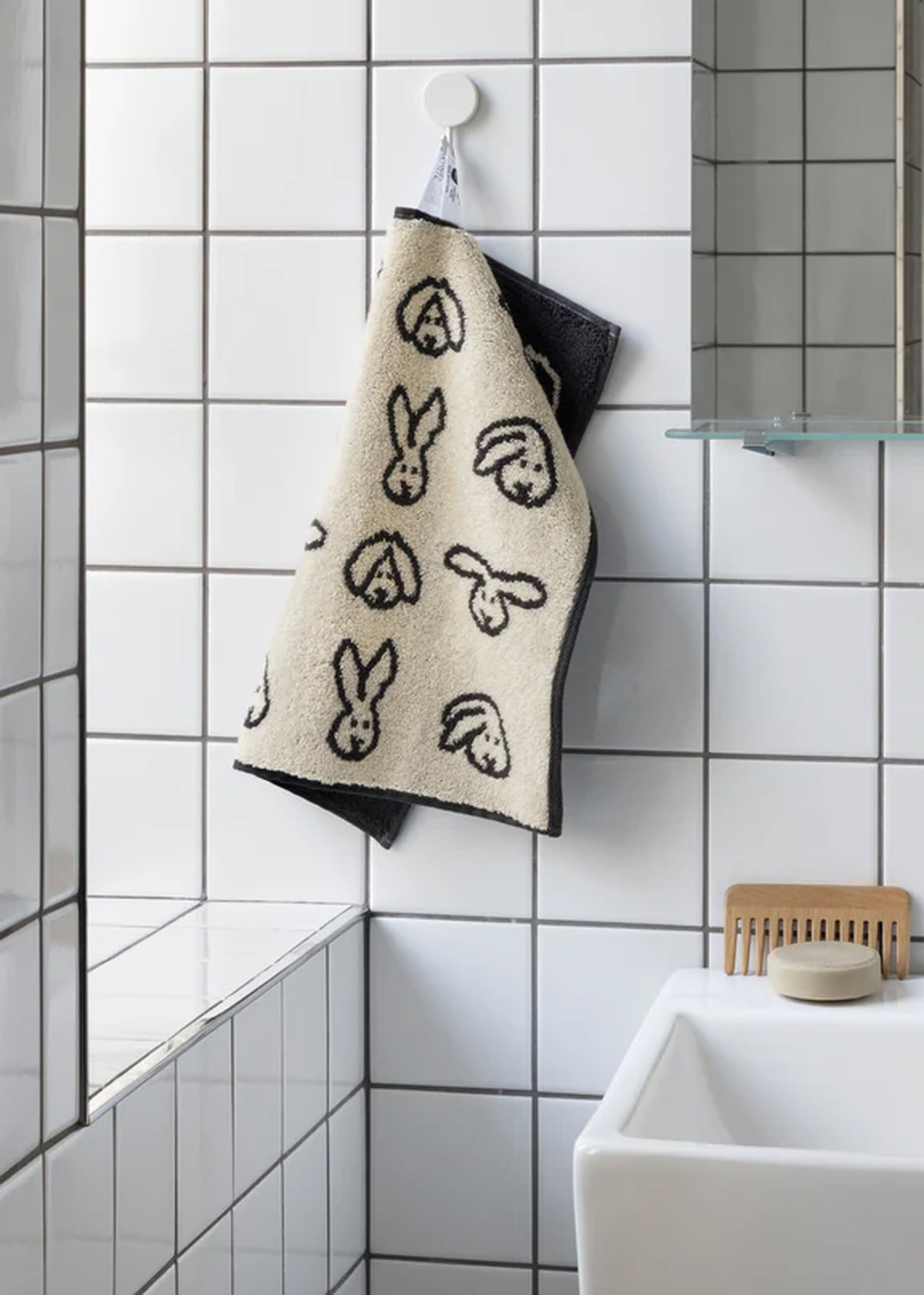 The WARMGREY TAIL Bunny Bunny Hand Towel in cream and black, featuring abstract bunny faces, hangs on a white tile wall beside a sink, comb, and glass shelf in a modern bathroom.