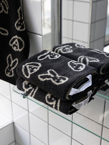 WARMGREY TAIL Bunny Bunny Hand Towels, featuring black soft cotton and white bunny face patterns, are neatly folded on a glass bathroom shelf, with a matching WARMGREY TAIL hand towel hanging nearby.