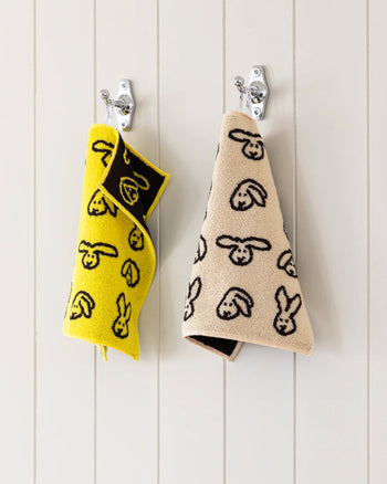 Two WARMGREY TAIL Bunny Bunny Hand Towels, one yellow and one beige with cartoon faces, hang on silver hooks against a white paneled wall. Made from soft cotton towel fabric for extra comfort.
