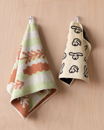 Two patterned hand towels hang on a light wooden wall. The left is a soft cotton towel with green, white, and brown stripes and leaf shapes; the right is the WARMGREY TAIL Bunny Bunny Hand Towel by WARMGREY TAIL, beige with playful black bunny faces.