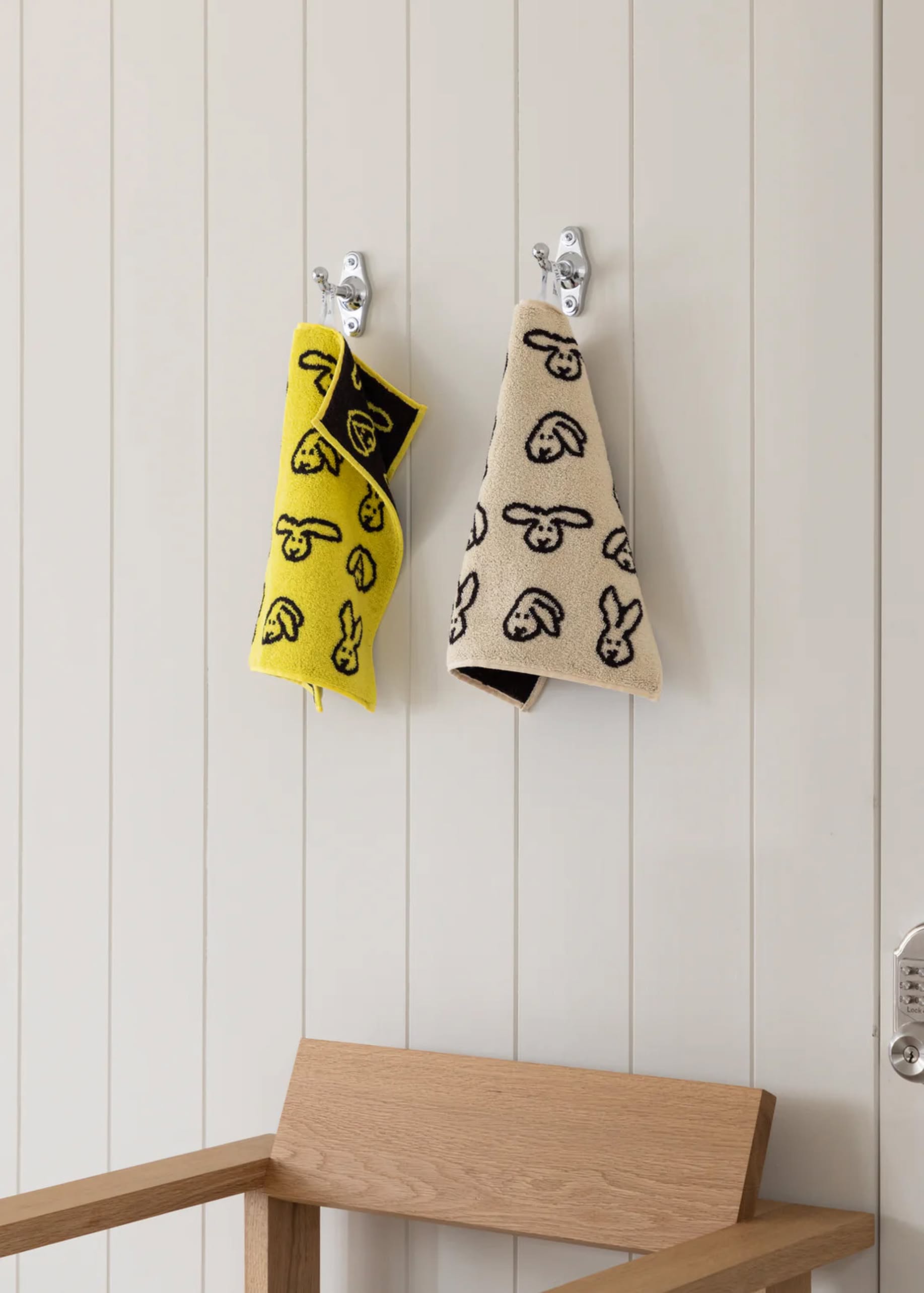 Two WARMGREY TAIL Bunny Bunny Hand Towels hang on wall hooks above a wooden chair. One is yellow, the other beige, both featuring black bunny faces. Light vertical paneling adds a charming backdrop.