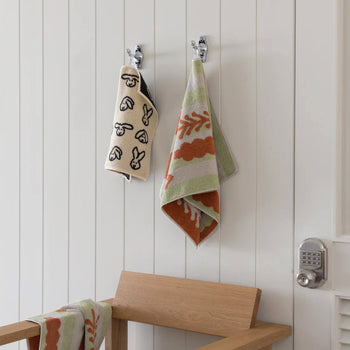 Two playful bathroom towels hang on hooks above a wooden bench—a WARMGREY TAIL Bunny Bunny Hand Towel with animal faces, and another with a nature-inspired print. A keypad door lock appears to the right on the white paneled wall.