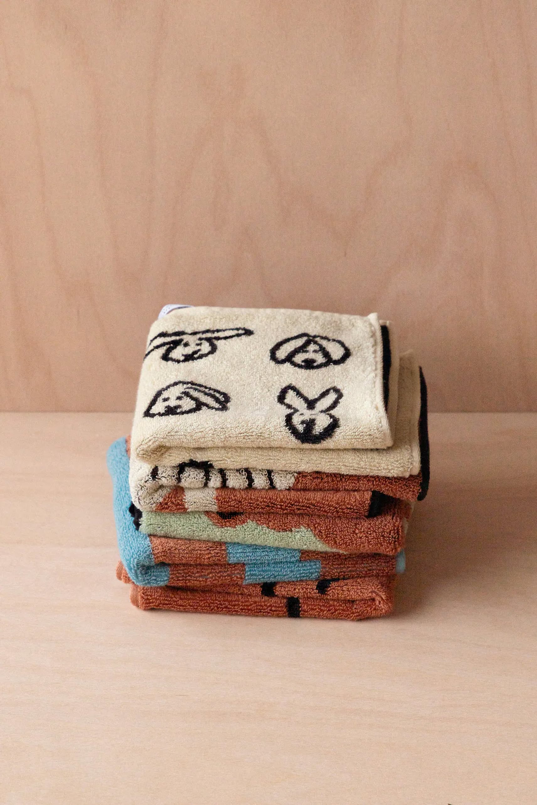 A stack of WARMGREY TAIL Bunny Bunny Hand Towels from WARMGREY TAIL in beige with black abstract animal face designs sits on a light wooden surface, joined by other soft cotton towels in brown, blue, and green tones.