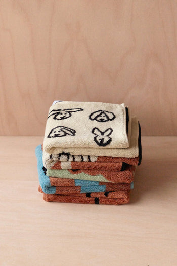 A stack of WARMGREY TAIL Bunny Bunny Hand Towels from WARMGREY TAIL in beige with black abstract animal face designs sits on a light wooden surface, joined by other soft cotton towels in brown, blue, and green tones.