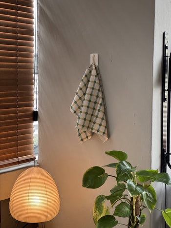 A WARMGREY TAIL Dot Check Kitchen Cloth 2 hangs on a white wall hook above a green plant. Nearby, a round lamp glows beside wooden blinds—adding warmth and charm to your kitchen with this stylish WARMGREY TAIL accessory.