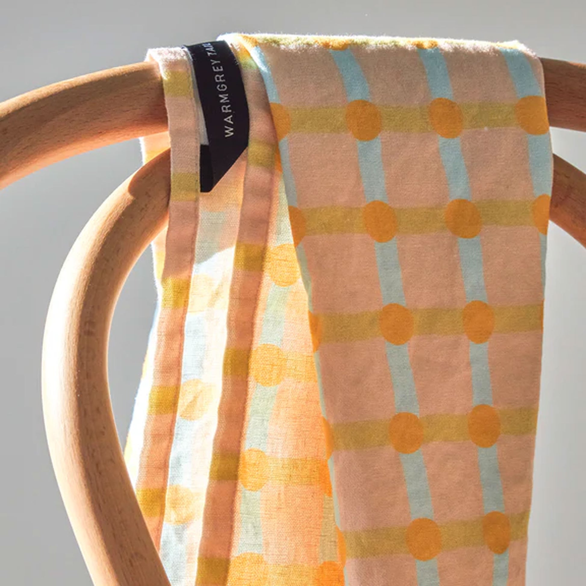 The WARMGREY TAIL Dot Check Kitchen Cloth 2, in soft cotton with orange dots and blue lines on beige, drapes over a curved wooden chair. The fabric features a black tag with the WARMGREY TAIL brand name in white.