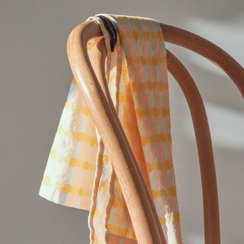 The WARMGREY TAIL Dot Check Kitchen Cloth 2, featuring yellow and orange patterns, is draped over a curved wooden chair backrest, bathed in gentle natural light.