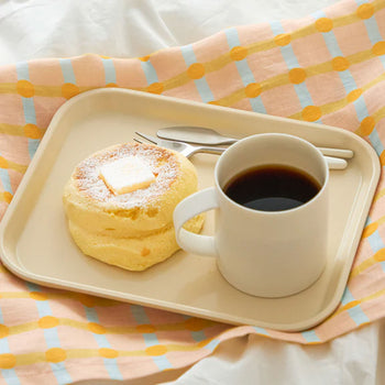 A beige tray with pancakes topped with butter and powdered sugar, a fork and knife, and a white mug of black coffee rests on the WARMGREY TAIL Dot Check Kitchen Cloth 2 by WARMGREY TAIL in pastel checkered patterns.