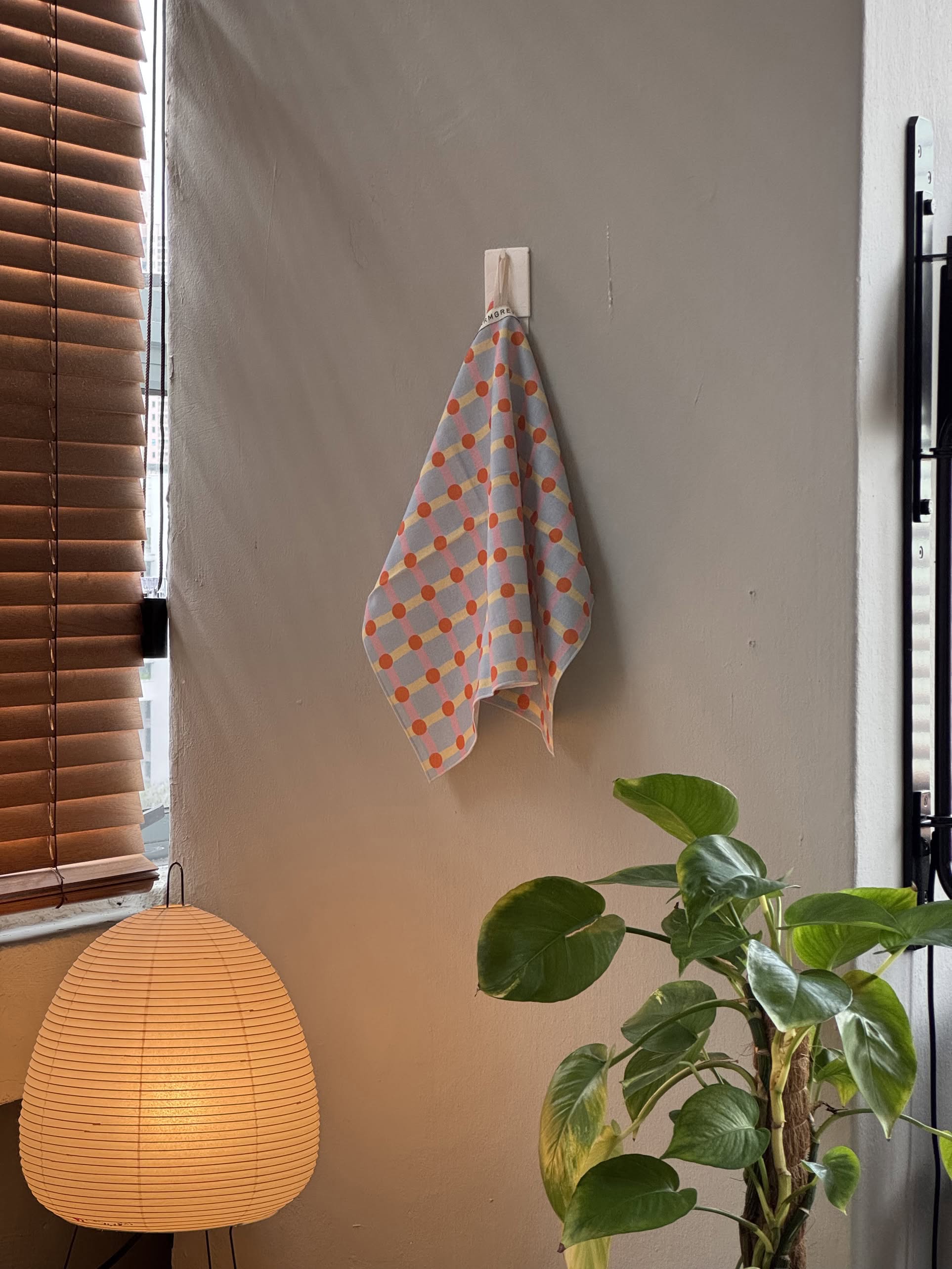 The WARMGREY TAIL Dot Check Kitchen Cloth 2, featuring orange and blue squares, hangs by a window with wooden blinds—a stylish accessory from WARMGREY TAIL to brighten any kitchen.