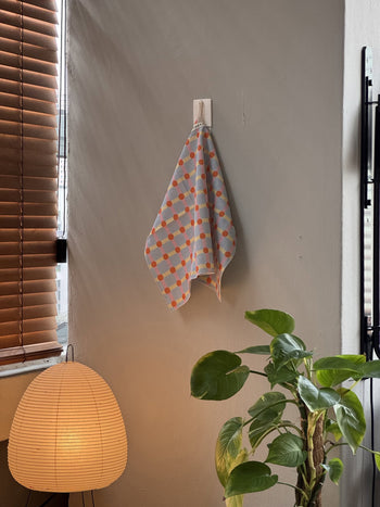 The WARMGREY TAIL Dot Check Kitchen Cloth 2, featuring orange and blue squares, hangs by a window with wooden blinds—a stylish accessory from WARMGREY TAIL to brighten any kitchen.