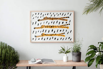 A wooden table with potted plants and an open book sits against a white wall, topped by the WARMGREY TAIL “Run Cheetah Run!” Poster (30x40), a minimalist artwork of three abstract cheetahs by WARMGREY TAIL.