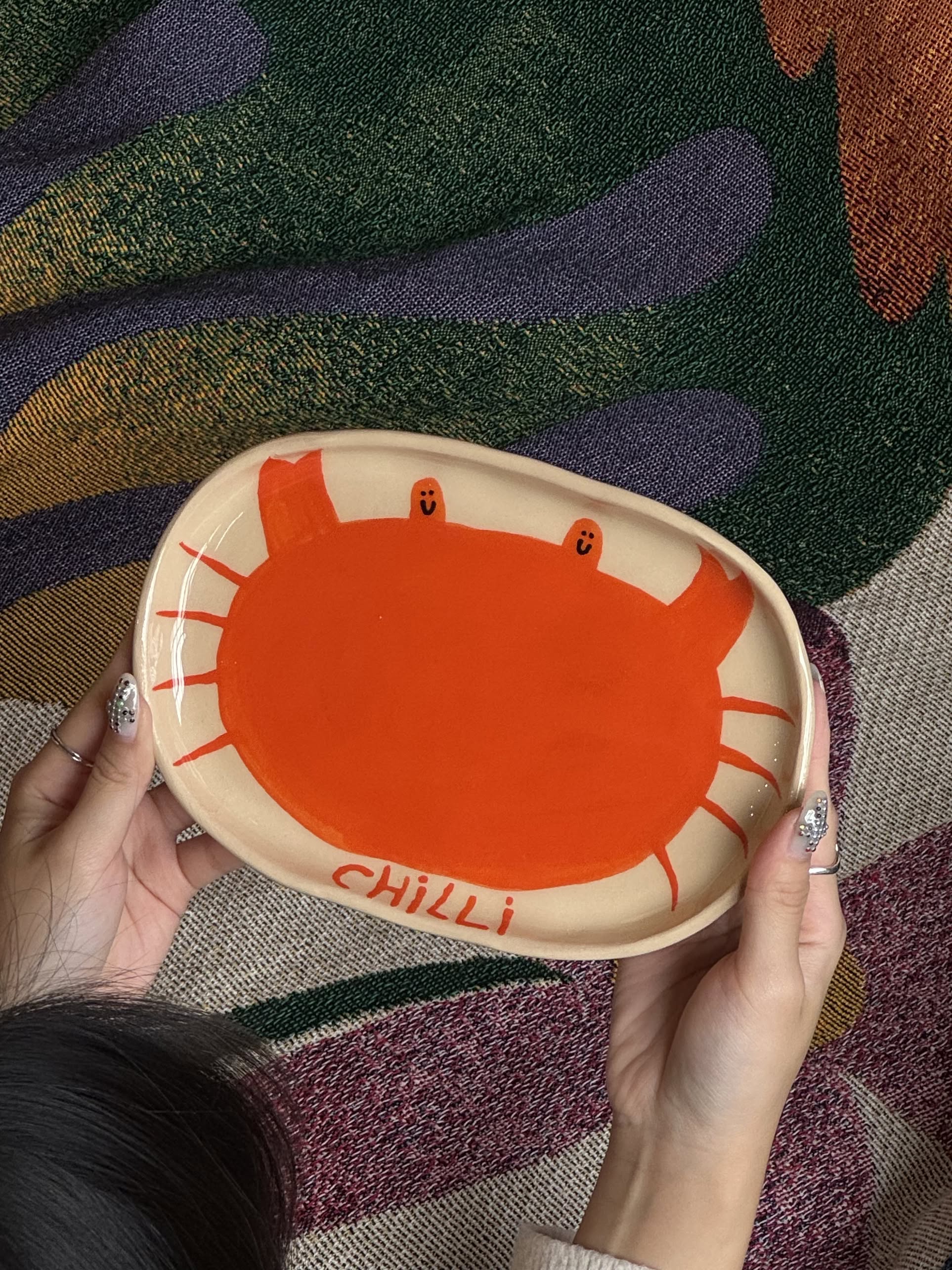 A person holds the Wobbly Studio Stacked Store Exclusive Happy Things Plate – Happy Chilli Crab, a handmade earthenware plate with a red crab and “CHILLI” design, set against a colorful abstract patterned fabric background. Perfect for Singapore Chilli Crab fans!.