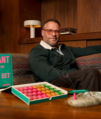 A man with glasses sits on a patterned sofa beside the ripple+ Houseplant Ash & Scent Set, with one cone burning in the marble ashtray. Wooden walls and a retro lamp complete the room’s cozy vibe.