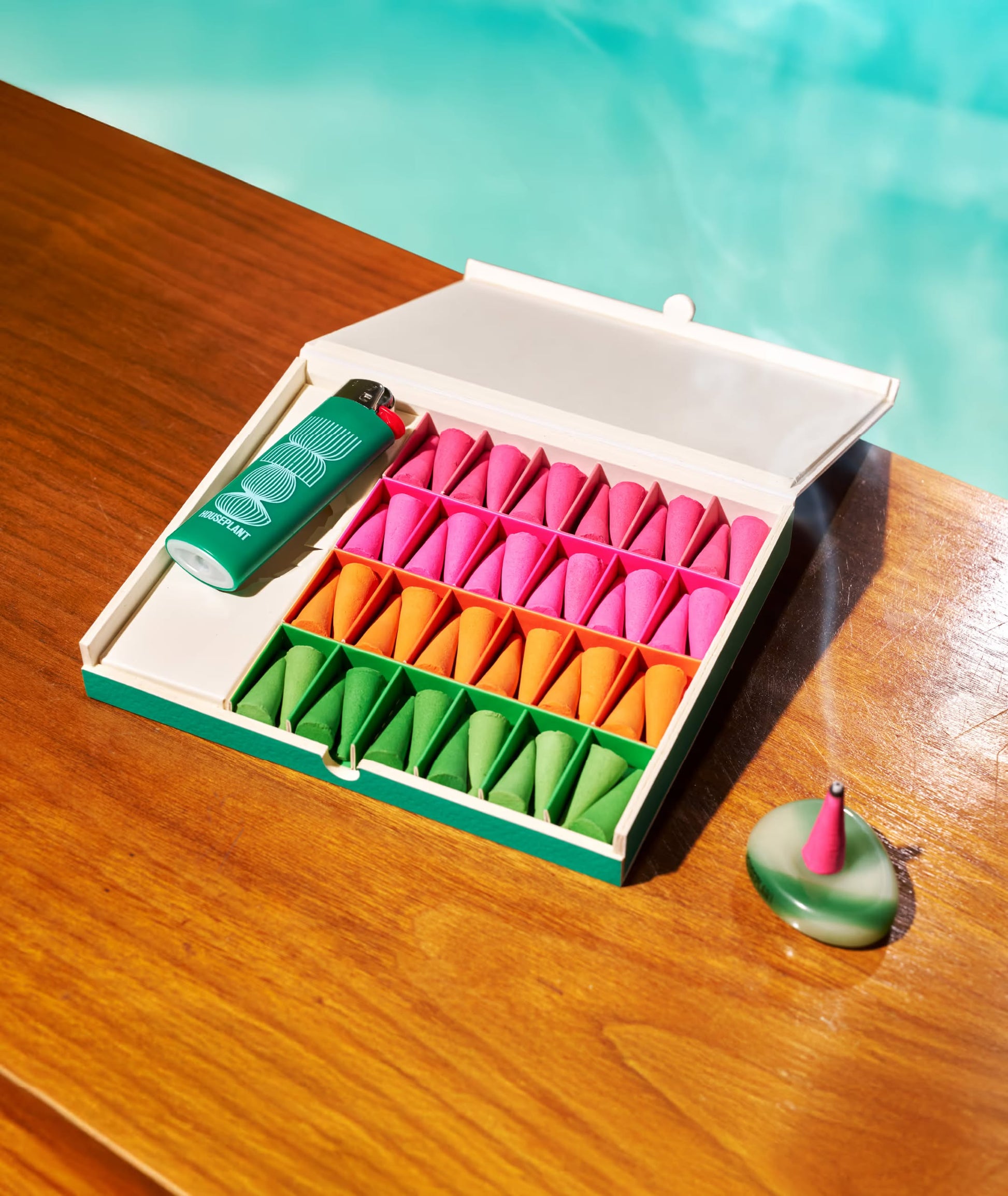 The ripple+ Houseplant Incense Droplets Sampler Set, featuring hand-rolled incense and a green lighter, sits on a wooden surface by a turquoise pool beside a burning cone—bringing calm to your space.