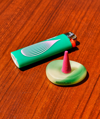 A ripple+ Houseplant Incense Droplets Sampler Set cone sits on a pale green holder labeled HOUSEPLANT, beside a green lighter with pink and white design, both on a reddish wooden surface.