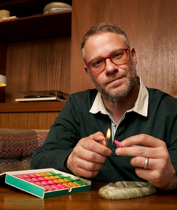 A man with short hair and glasses lights a colorful incense cone from the ripple+ Houseplant Incense Droplets Sampler Set at a wooden table, next to a smooth stone, in a retro room with wood-paneled walls.