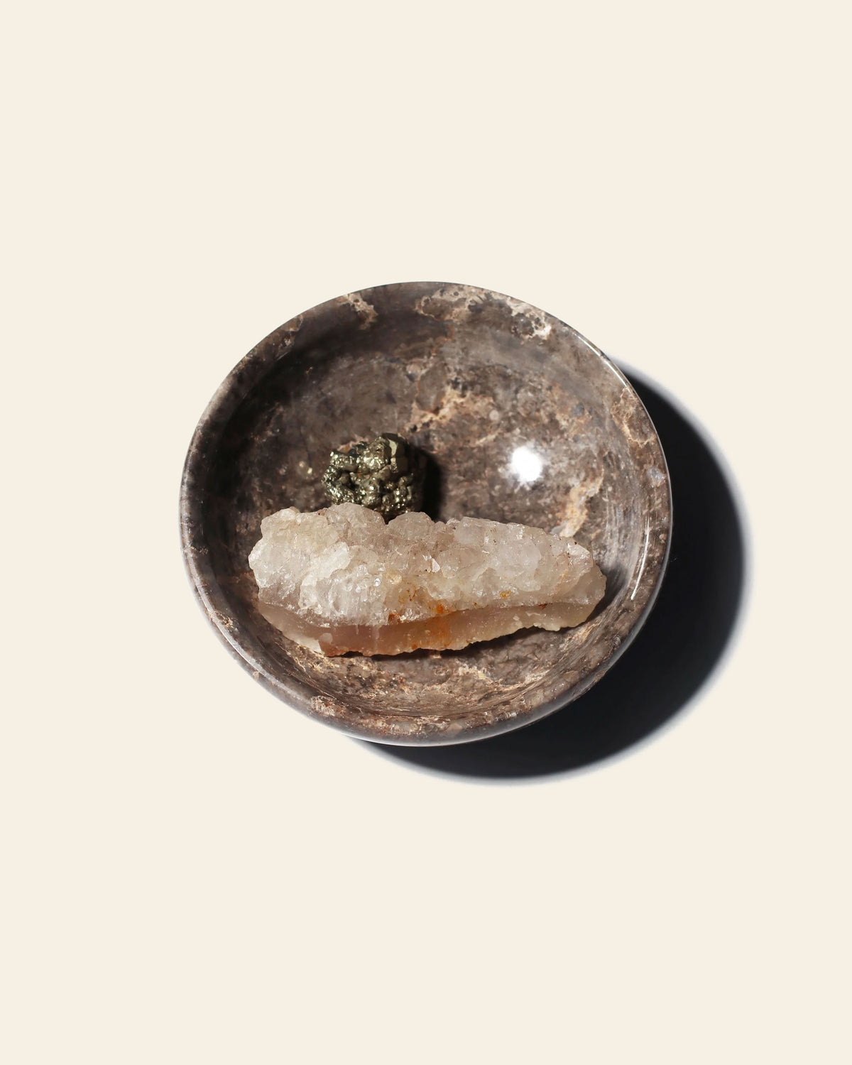 A small, round, dark stone stillgoods Luna Dish containing a clear crystalline shard and a cluster of metallic pyrite.